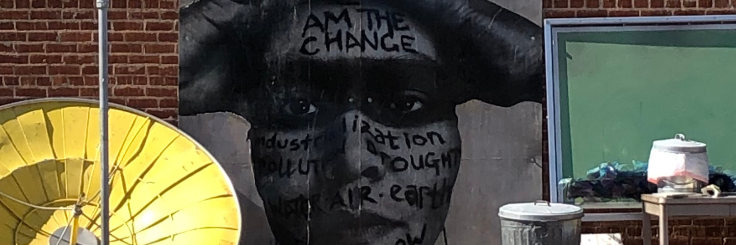 Face on large board with text “Am the Change. Industrialization, Pollution, Drought. Water, Air, Earth” written over face.